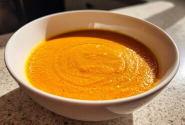 Close-up of a white bowl filled with vibrant orange Vegan Creamy Roasted Carrot Ginger Soup.