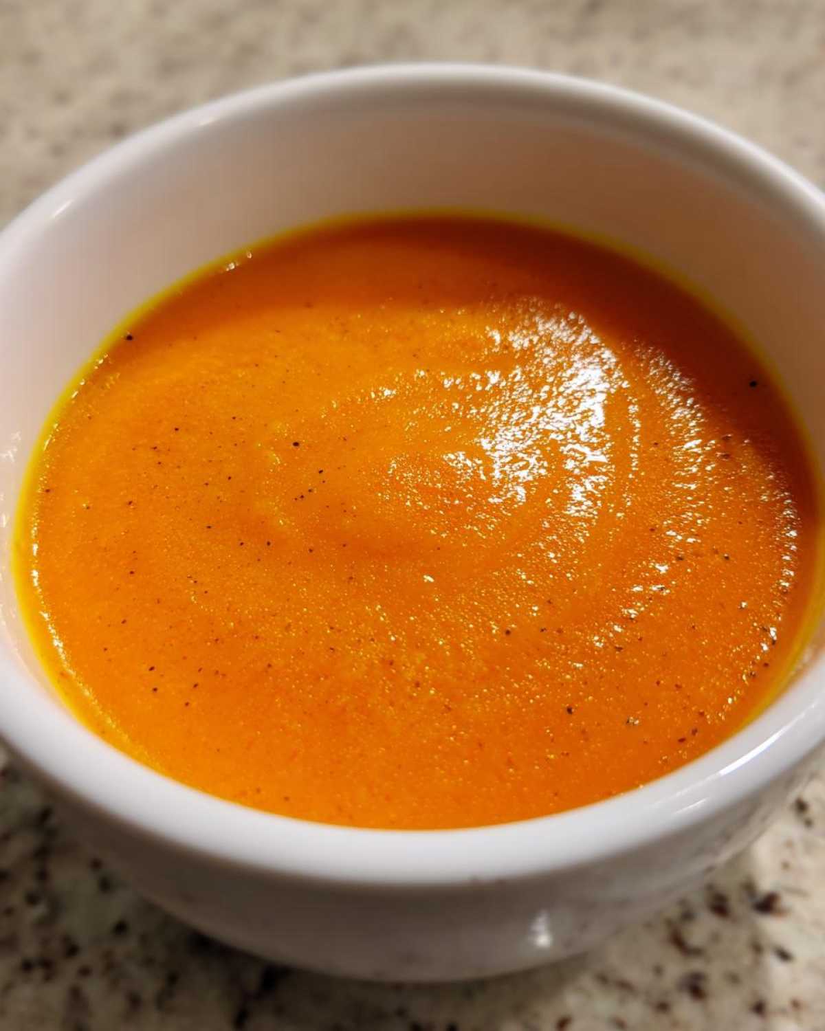 A close-up of vibrant orange Vegan Creamy Roasted Carrot Ginger Soup in a white bowl, lightly seasoned with pepper flecks.