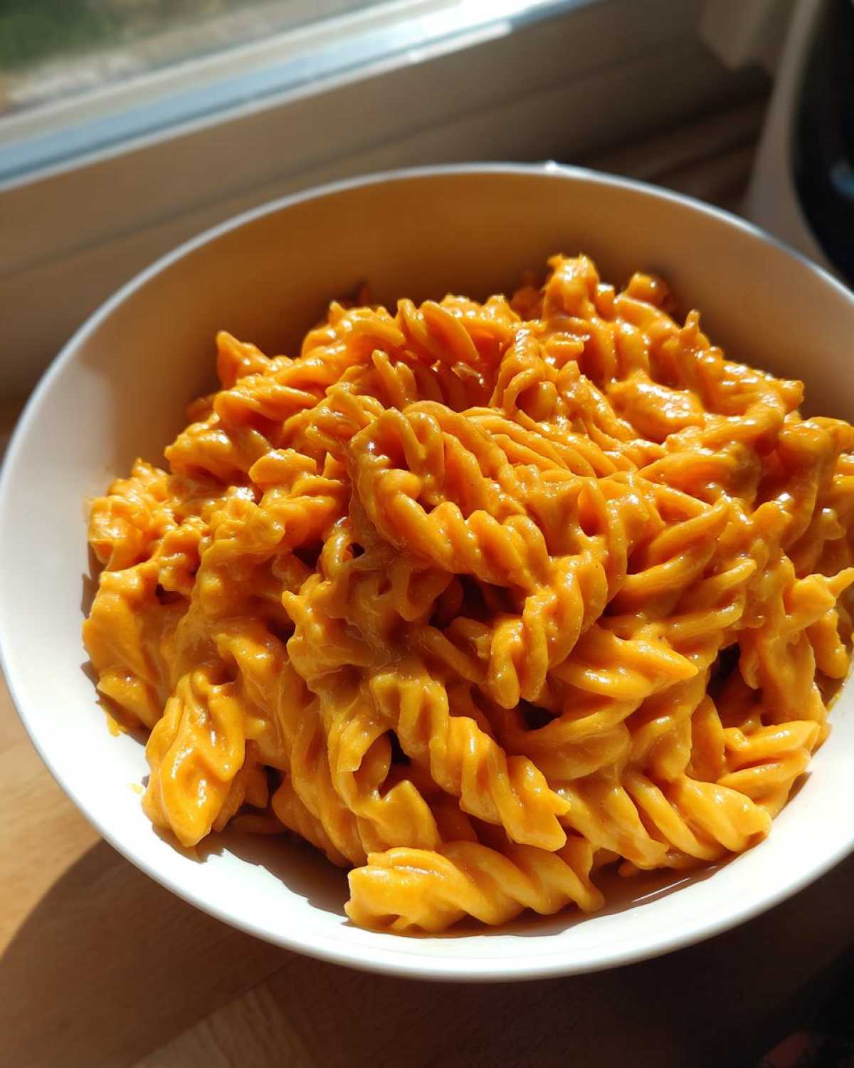 A white bowl filled with rotini pasta coated in bright orange Vegan Creamy Sweet Potato Mac Sauce, lit by sunlight.