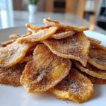 A close-up pile of crispy Air Fryer Cinnamon Sugar Banana Chips dusted heavily with sugar and cinnamon on a white plate.