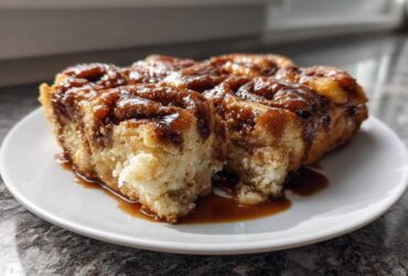 A close-up of several squares of moist Amish Cinnamon Flop covered in rich, dark caramel glaze on a white plate.