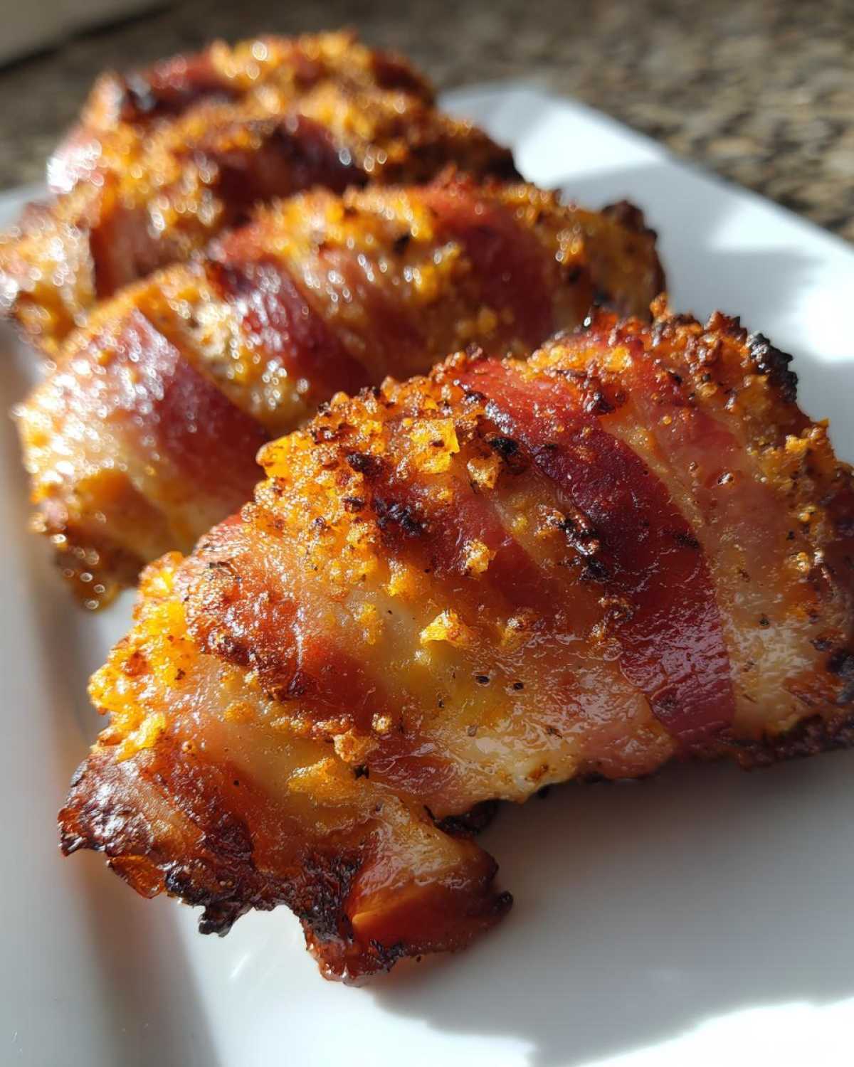 Close-up of crispy, baked Bacon Wrapped Garlic Parmesan Chicken pieces lined up on a white plate.