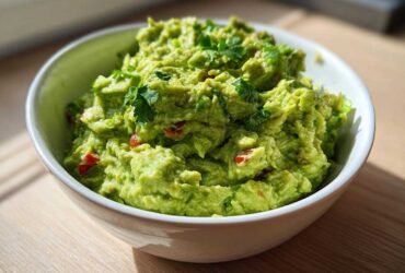 A close-up of chunky, bright green basic guacamole topped with cilantro in a white bowl.