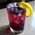 Close-up of a fizzy glass of Blueberry Lemonade garnished with fresh blueberries and a lemon slice.