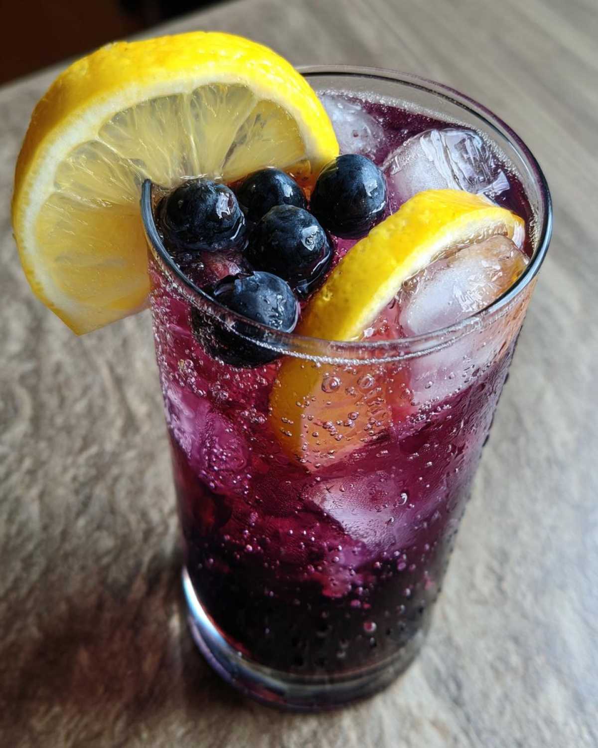 Close-up of a tall glass filled with sparkling Blueberry Lemonade, ice cubes, fresh blueberries, and lemon slices.