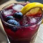 Close-up of a tall, sweating glass filled with vibrant Blueberry Lemonade, ice cubes, fresh blueberries, and a lemon slice.