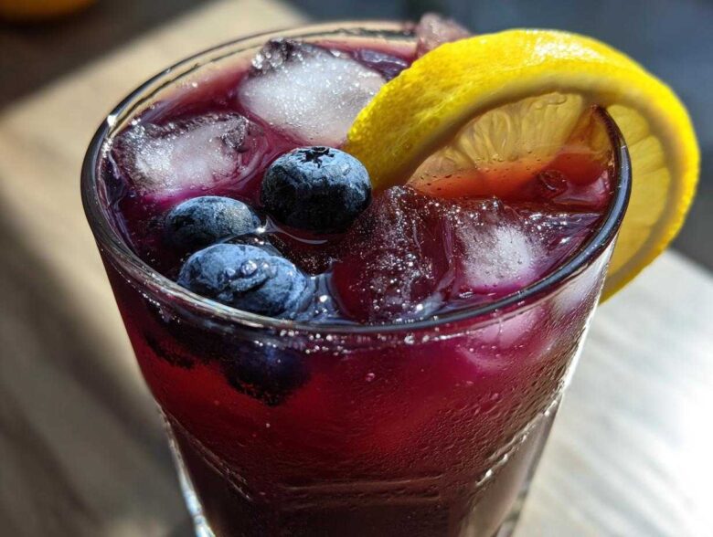 Close-up of a tall, sweating glass filled with vibrant Blueberry Lemonade, ice cubes, fresh blueberries, and a lemon slice.