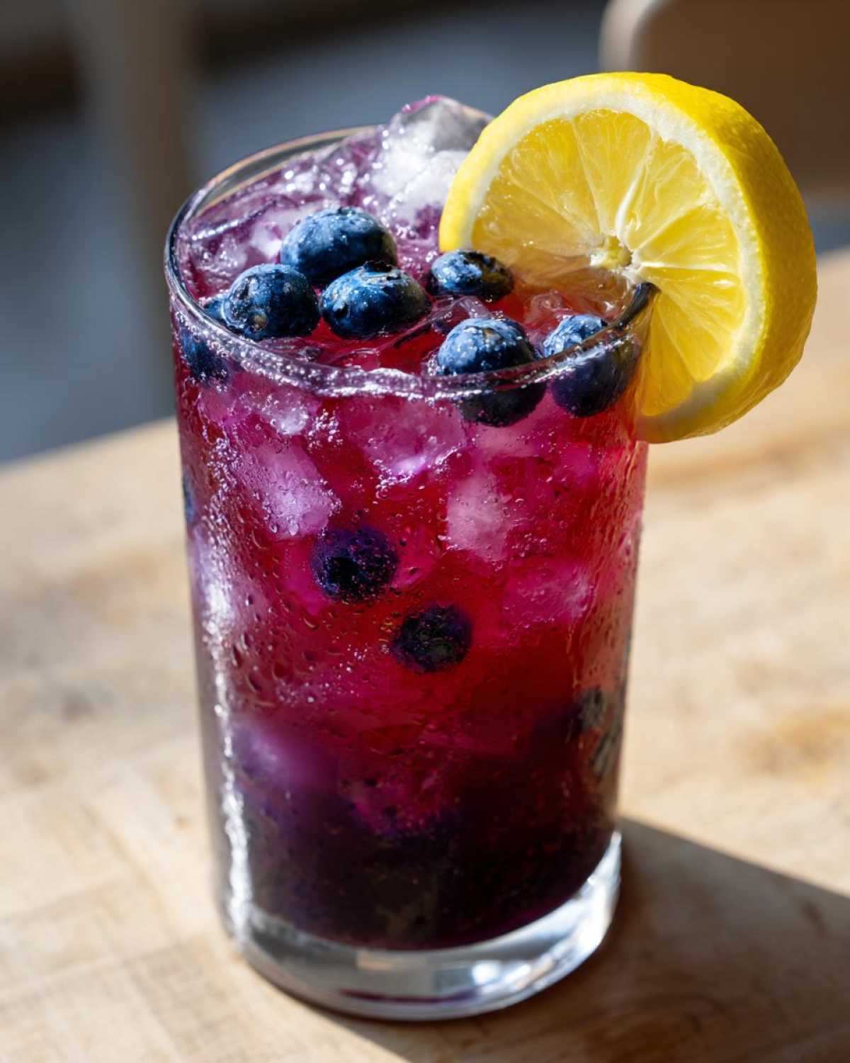 Close-up of a tall glass filled with vibrant purple Blueberry Lemonade, ice cubes, fresh blueberries, and a lemon slice garnish.