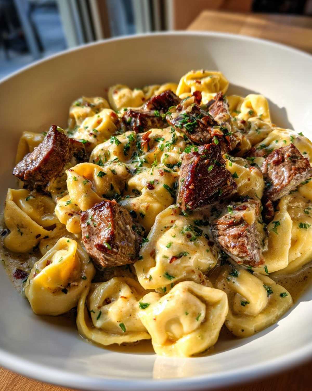 A close-up, bright photo of Cracked Garlic Steak Tortellini tossed in a creamy, garlicky sauce and topped with chunks of seared steak and parsley.
