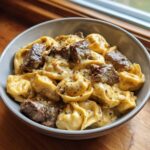 A close-up of a bowl filled with creamy Cracked Garlic Steak Tortellini, topped with chunks of seared steak and cracked pepper.