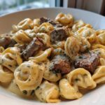 Close-up of a white bowl filled with Cracked Garlic Steak Tortellini coated in a creamy sauce with chunks of steak.