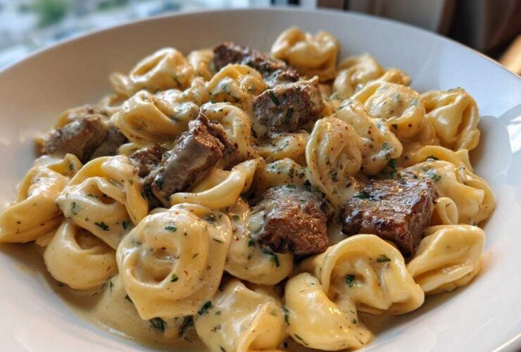 Close-up of a white bowl filled with Cracked Garlic Steak Tortellini coated in a creamy sauce with chunks of steak.