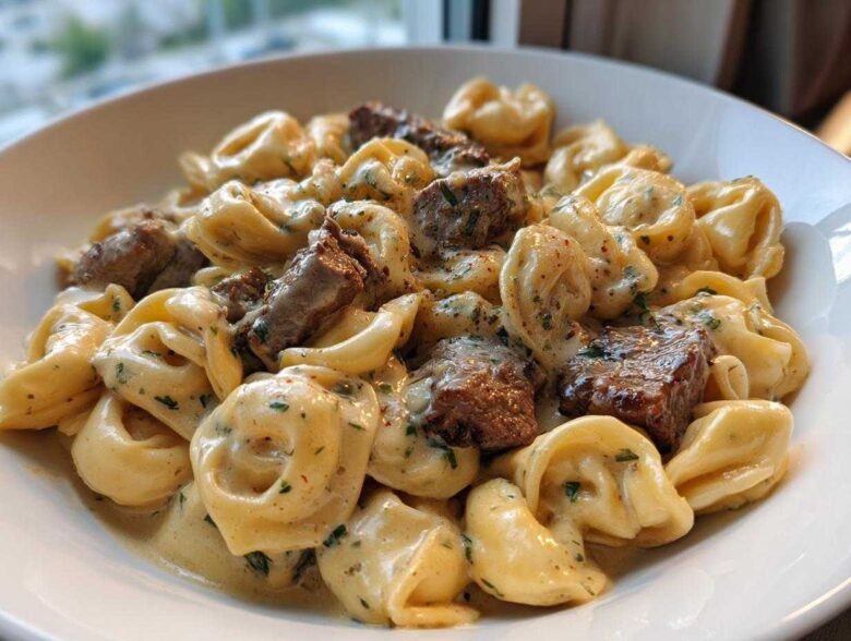 Close-up of a white bowl filled with Cracked Garlic Steak Tortellini coated in a creamy sauce with chunks of steak.