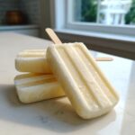 Three creamy, pale yellow Banana Coconut Popsicles stacked on a white counter with a wooden stick visible.