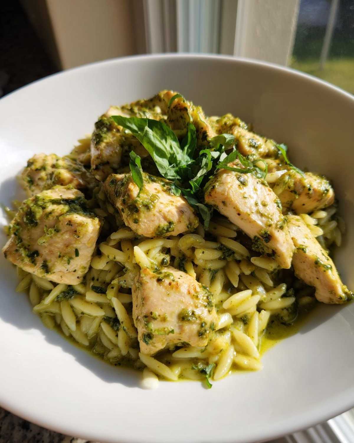 A white bowl filled with creamy pesto chicken pieces served over orzo pasta, garnished with fresh basil.