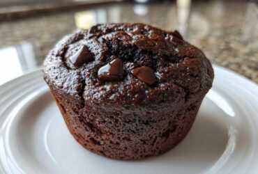A close-up of a rich, dark Double Chocolate Muffin topped with glossy, melted chocolate chips on a white plate.