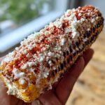 A hand holding a grilled ear of Elote with Cotija Cheese and Lime, generously topped with white cheese and red chili powder.