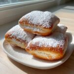 A stack of four golden brown French Beignets generously dusted with white powdered sugar on a white plate.