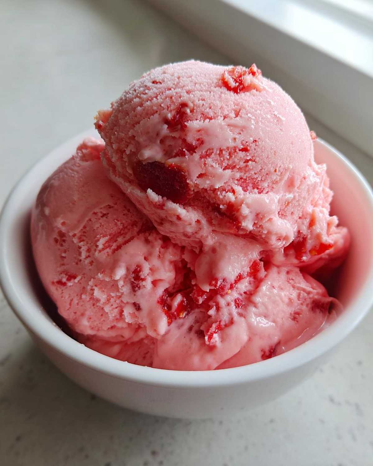 Two scoops of pink Fresh Strawberry Dream Ice Cream with visible strawberry pieces served in a white bowl.