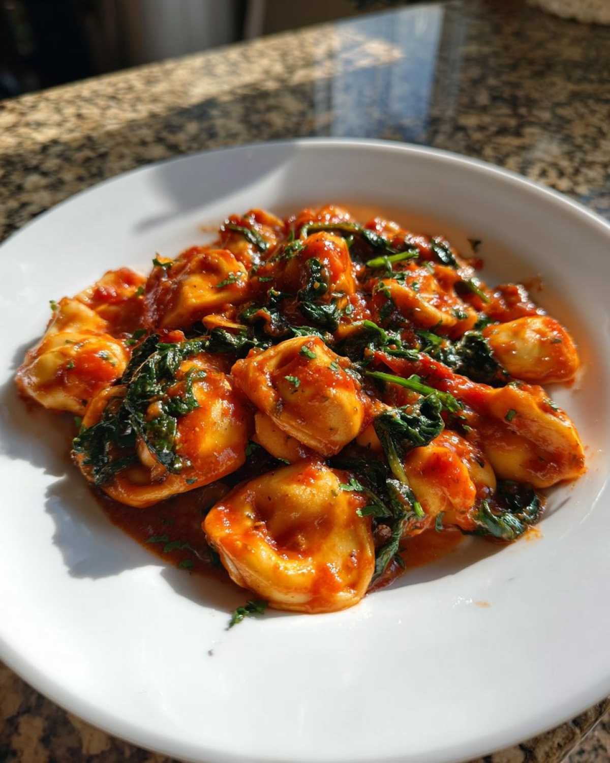 Close-up of a white plate filled with Garlic and Herb Tomato Tortellini mixed with wilted spinach in a rich tomato sauce.