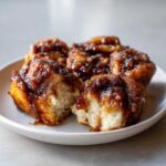 Four sticky, gooey Amish Cinnamon Flop rolls on a white plate, one broken open showing the soft interior.