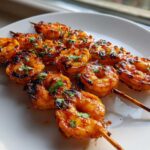 Close-up of several Grilled Sweet Chili Bang Bang Shrimp Skewers glazed with sauce and garnished with parsley on a white plate.