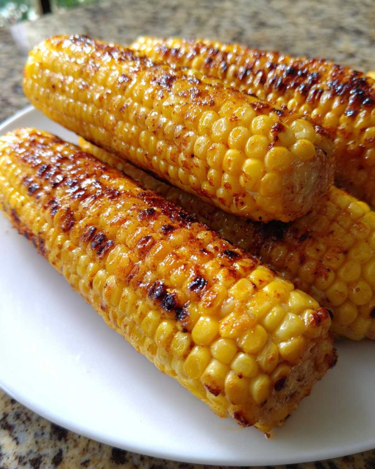 Close-up of several ears of golden yellow Grilled Sweet Corn on the Cob with visible char marks on a white plate.
