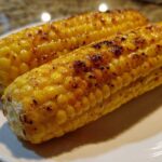 Close-up of two perfectly Grilled Sweet Corn on the Cob cobs glistening on a white plate with visible char marks.