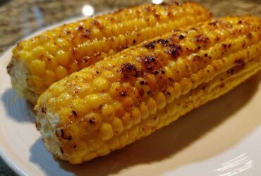 Close-up of two perfectly Grilled Sweet Corn on the Cob cobs glistening on a white plate with visible char marks.