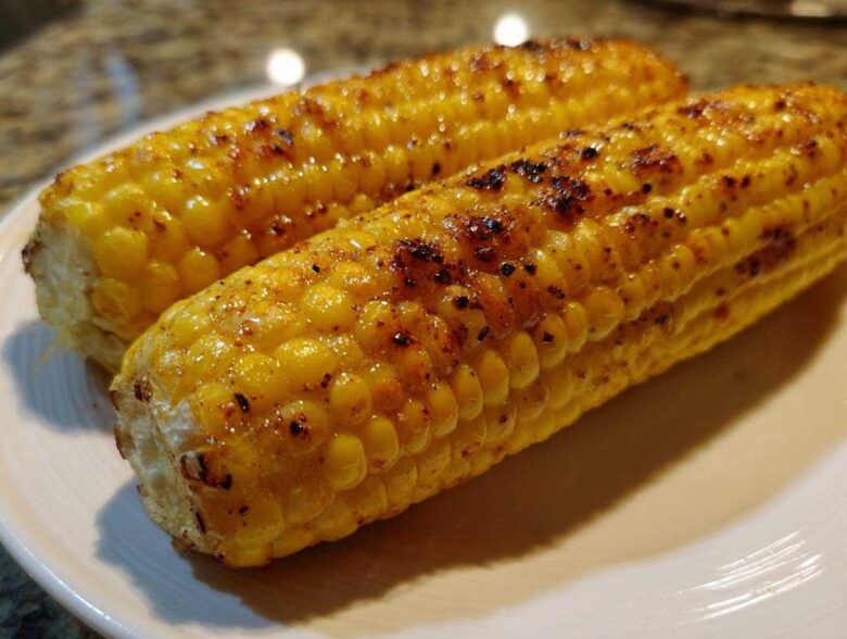 Close-up of two perfectly Grilled Sweet Corn on the Cob cobs glistening on a white plate with visible char marks.