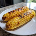 Two ears of glistening, perfectly Grilled Sweet Corn on the Cob with visible char marks, served on a white plate.
