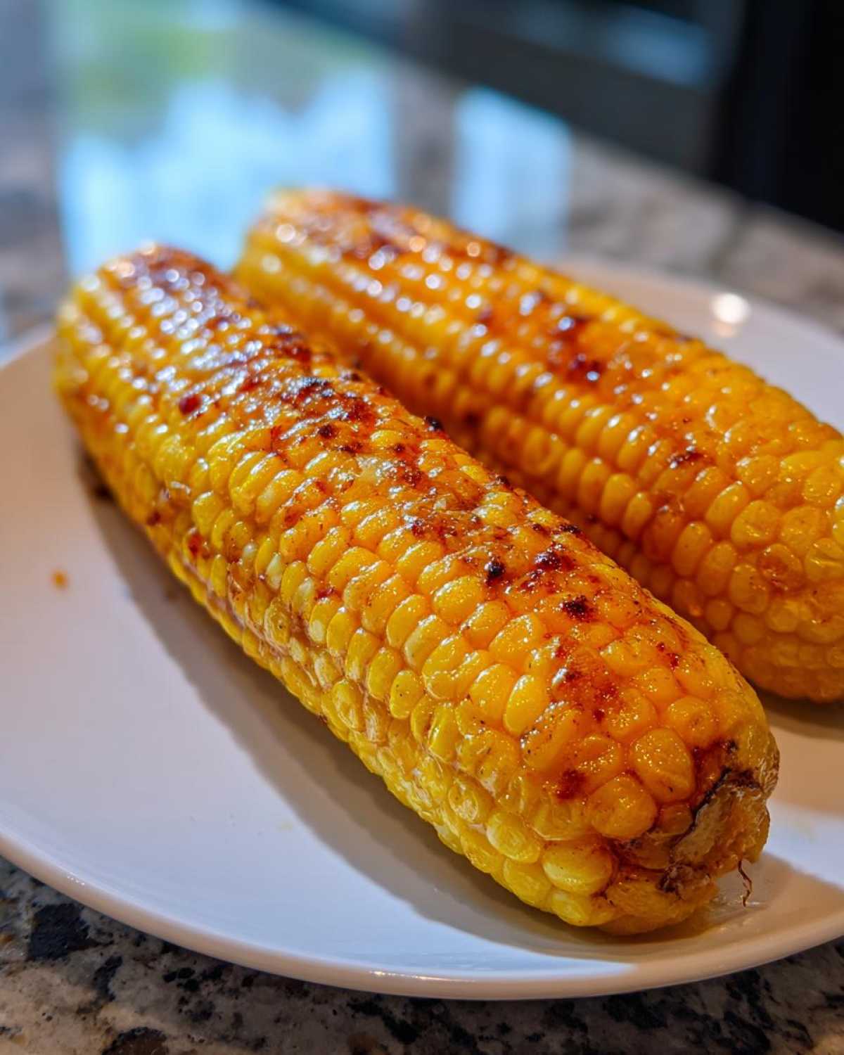 Two ears of golden yellow Grilled Sweet Corn on the Cob, glistening with seasoning, resting on a white plate.