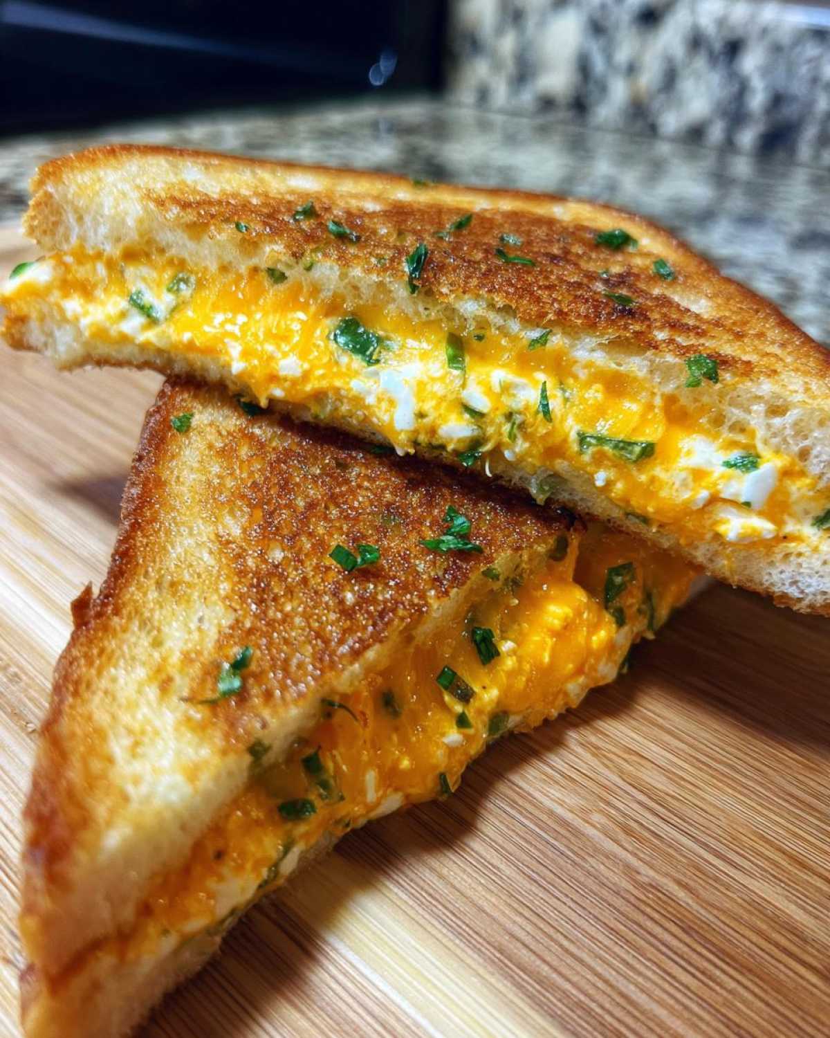 Two halves of a golden-brown Jalapeño Popper Grilled Cheese Sandwich stacked on a wooden board, showing melted orange and white cheese filling.