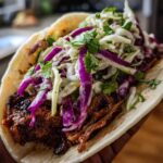 Close-up of a hand holding a soft tortilla filled with saucy mushroom filling and topped with purple and white cabbage slaw.