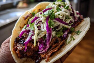 Close-up of a hand holding a soft tortilla filled with saucy mushroom filling and topped with purple and white cabbage slaw.