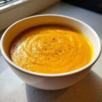 A close-up of a bowl of vibrant orange Roasted Apple Pumpkin Soup, swirled and topped with cracked black pepper.