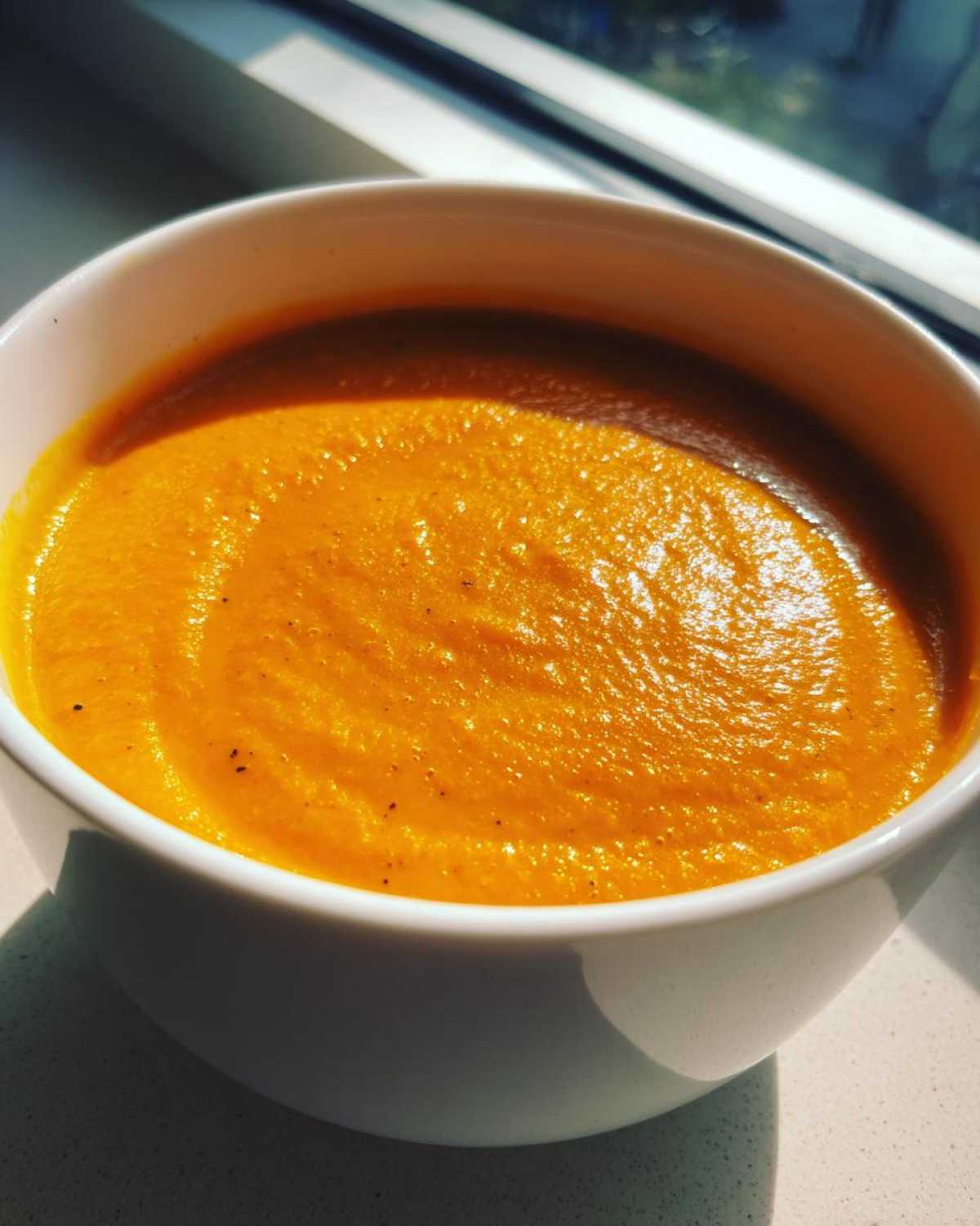 Close-up of a bowl filled with vibrant orange Roasted Apple Pumpkin Soup, highlighted by sunlight.