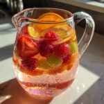 Close-up of a Ros Sangria Fruit Pitcher filled with sparkling pink liquid, oranges, strawberries, raspberries, and green grapes.