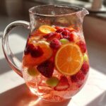 A clear glass Ros Sangria Fruit Pitcher filled with pink liquid, ice, orange slices, strawberries, raspberries, and green grapes.
