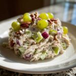 A mound of creamy The Chicken Salad mixed with celery, red onion, grapes, and pecans on a white plate.