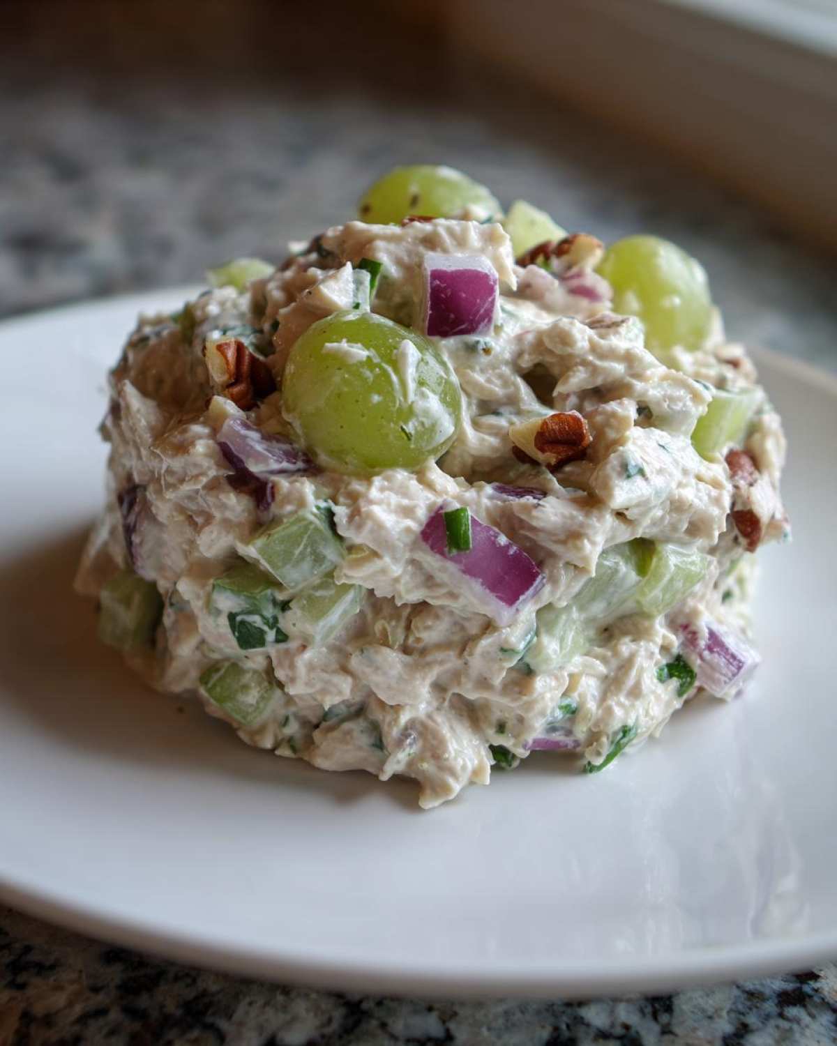 A mound of creamy The Chicken Salad mixed with green grapes, red onion, celery, and pecans on a white plate.