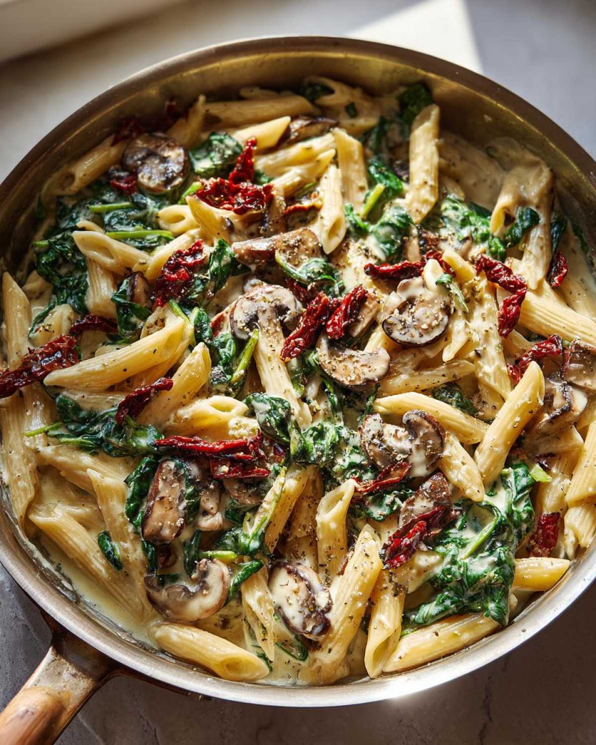 Overhead view of creamy Tuscan Mushroom Pasta Skillet with penne, mushrooms, spinach, and sun-dried tomatoes in a stainless steel skillet.