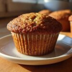 A close-up of a golden-brown Vanilla Chai Muffin sitting on a small white plate, with more muffins blurred in the background.