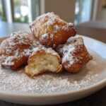 Close-up of four fluffy Vanilla French Beignets dusted heavily with powdered sugar on a light plate.