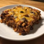 A square serving of cheesy Black Bean and Rice Bake topped with melted cheddar cheese on a white plate.