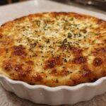 Close-up of a golden-brown, bubbly Creamy Baked Pickle Dip topped with herbs in a white fluted baking dish.