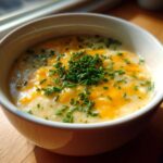 A close-up of a white bowl filled with rich Creamy Potato Soup, topped with melted cheddar cheese and fresh chopped chives.