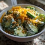 A white bowl filled with creamy Cucumber Ranch Crack Salad, topped with shredded cheddar cheese and cracker crumbs.