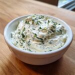 A close-up of a white bowl filled with thick, creamy Homemade Tzatziki Sauce mixed with visible green herbs.