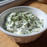 A close-up of creamy Homemade Tzatziki Sauce topped with fresh dill in a white bowl.